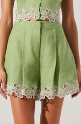 A woman is wearing a light green sleeveless top with decorative white floral embroidery along the hem and button area. The matching high-waisted shorts are also light green with the same white embroidered floral design along the hem, featuring intricate patterns of leaves and flowers.